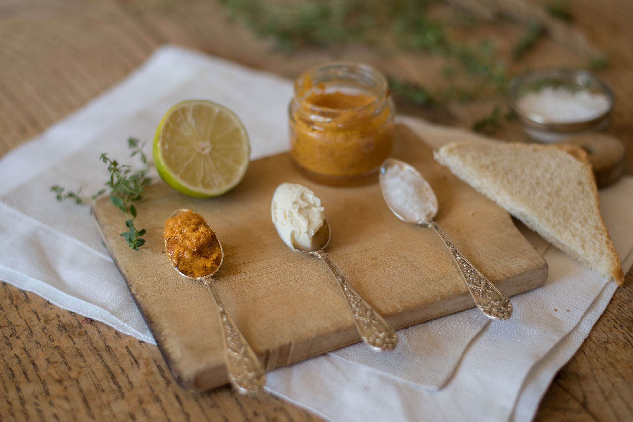 Drei Löffel mit verschiedenen Aufstrichen auf einem Holzbrett, daneben eine halbe Limette und Toast.