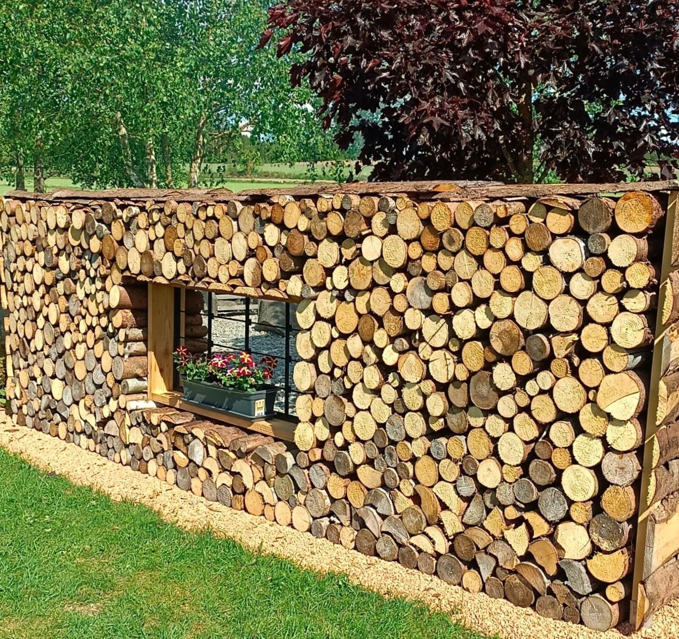 Holzwand mit Fenster und Blumenkasten, umgeben von Bäumen und Rasen.