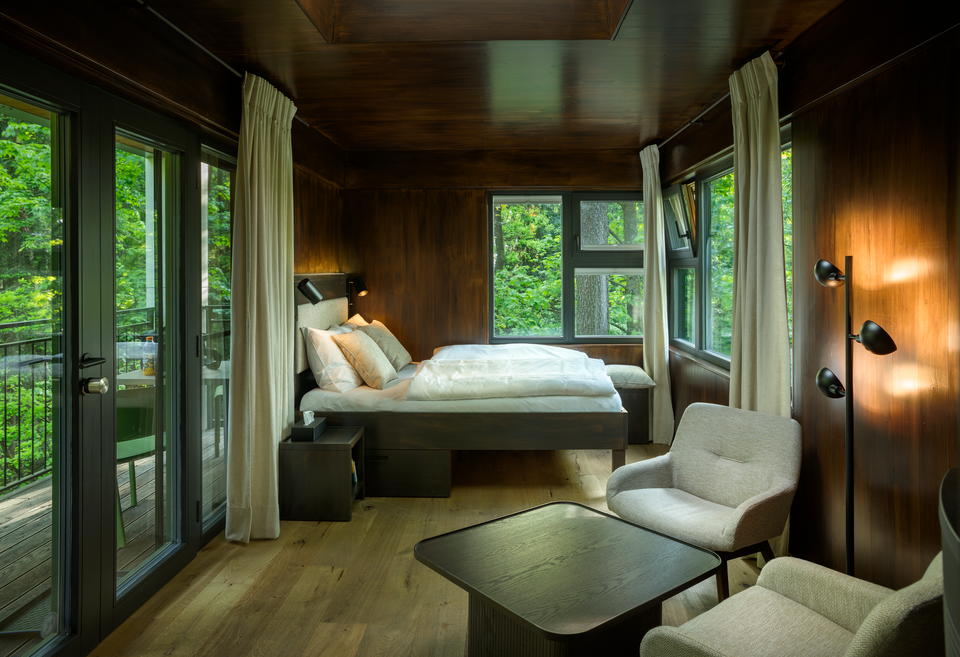 Interior view of a modern tree house with bed, armchairs and large windows with a view of the countryside.