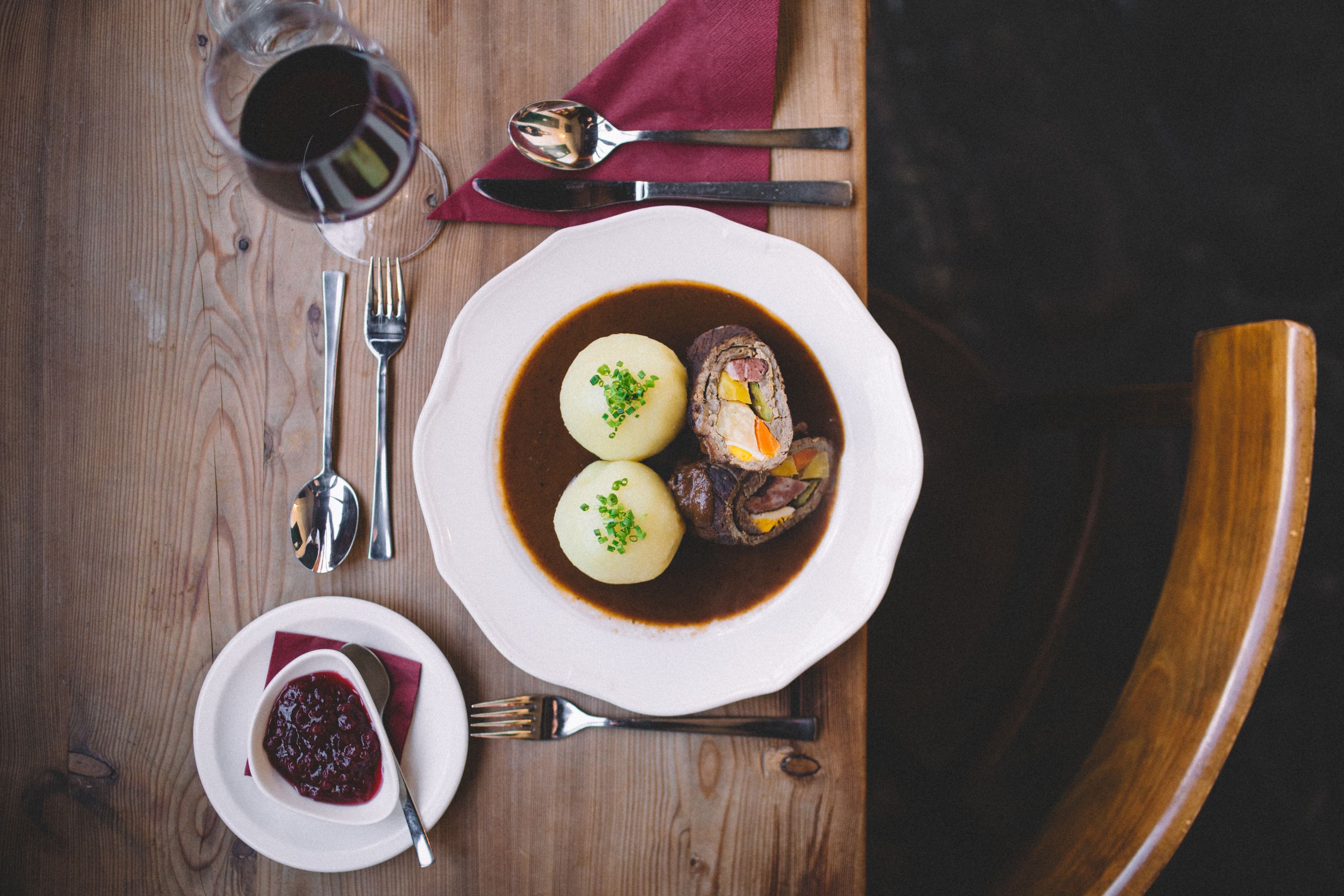 Ein Teller mit Rindsroulade und zwei Erdäpfelknödeln in Soße, daneben ein Glas Rotwein und Preiselbeeren auf einem Holztisch.