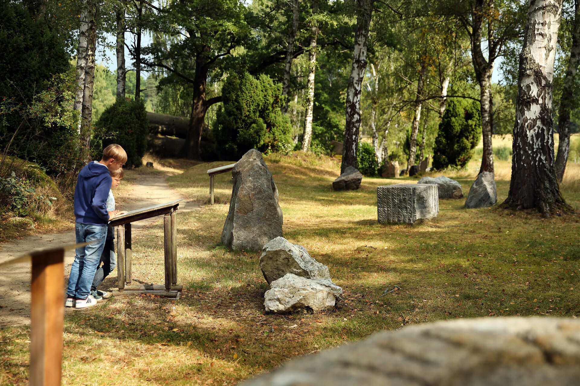 Zwei Kinder stehen an einer Informationstafel an einem Weg und lesen diese durch. Große Gesteinsblöcke liegen neben dem Weg. 