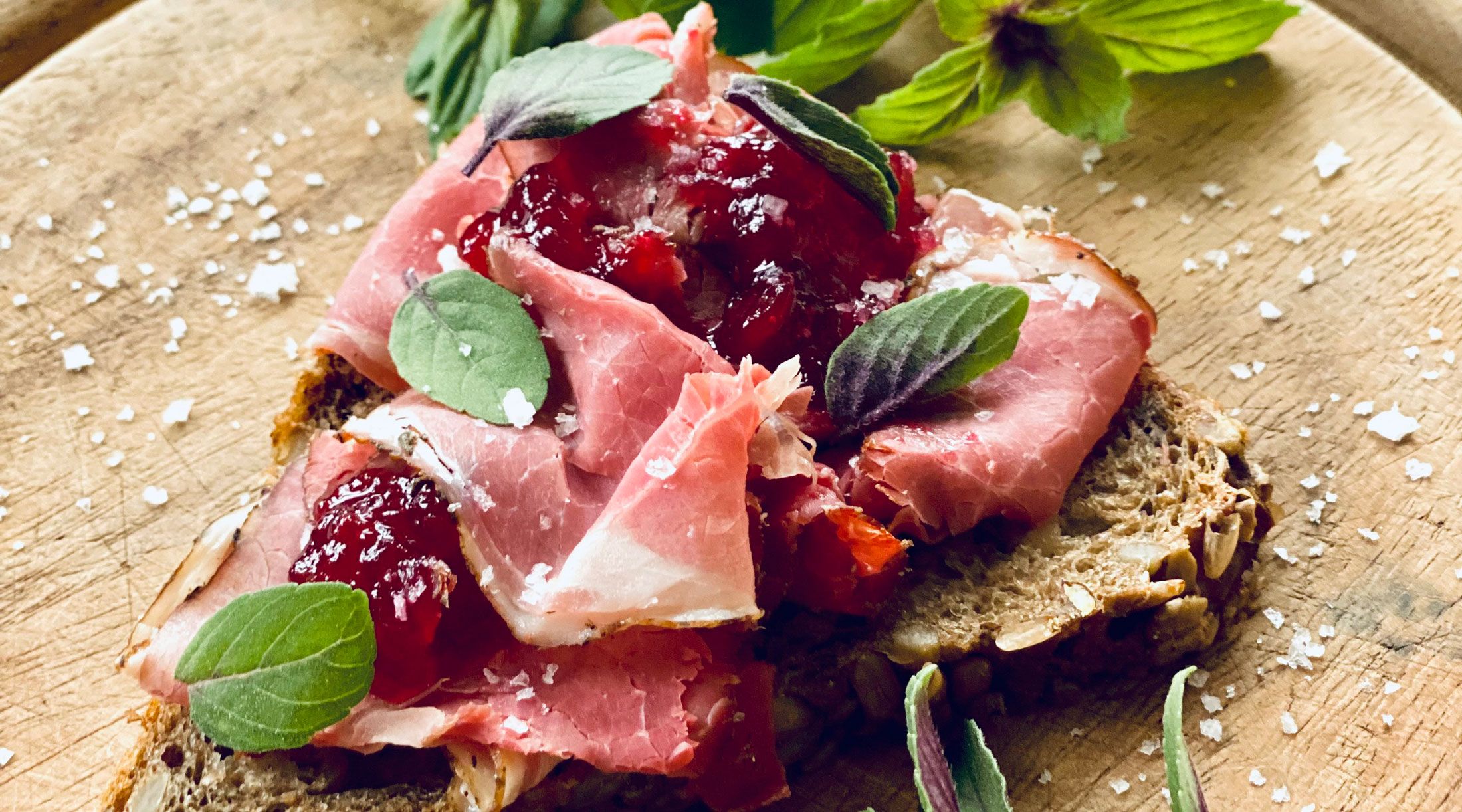 Nahaufnahme eines belegten Brotes mit Schinken, Preiselbeeren und Kräutern auf einem Holzbrett.