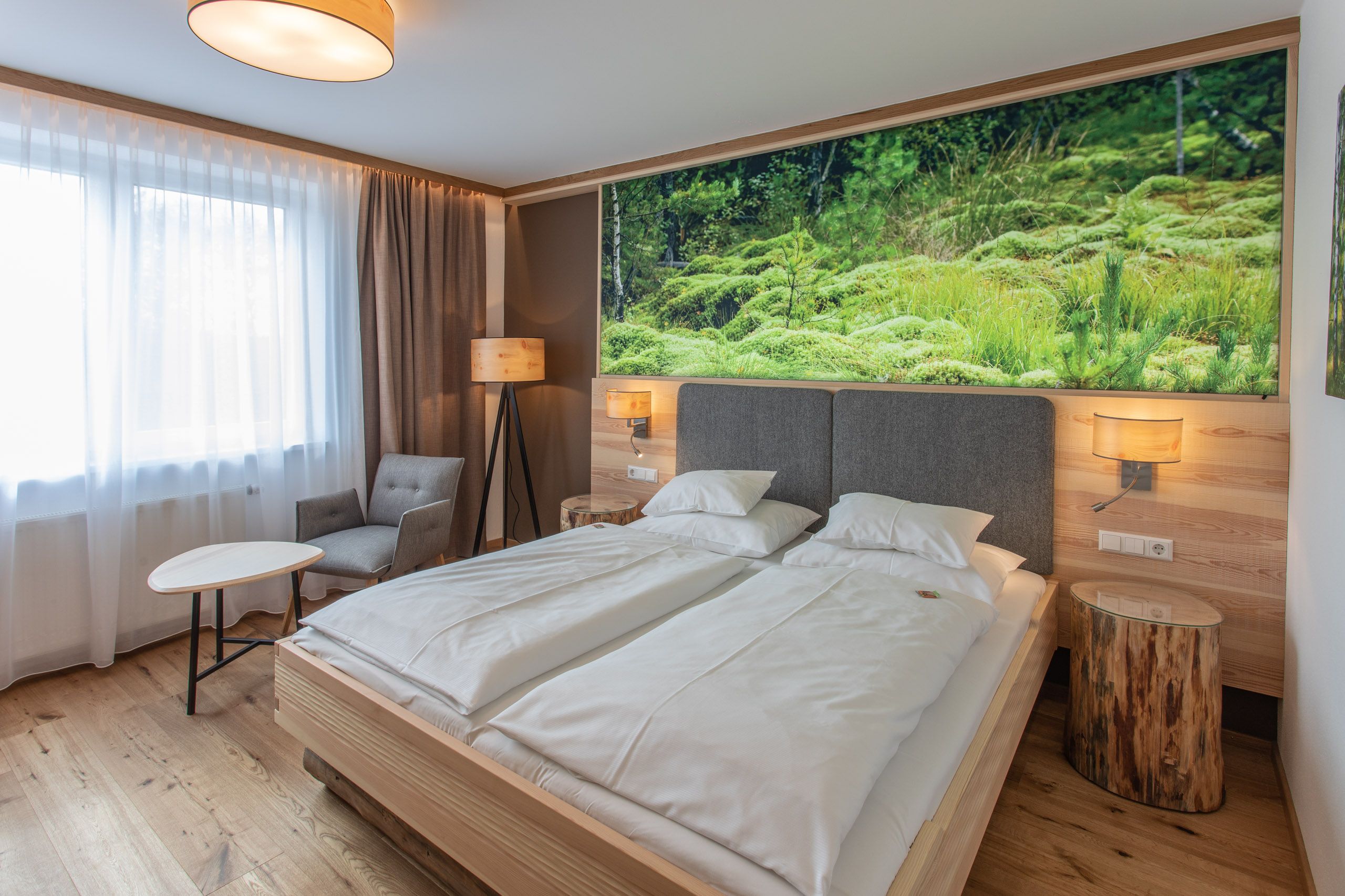 Modernes Hotelzimmer mit Doppelbett, Naturbild an der Wand, Sessel, Tisch und Stehlampe.