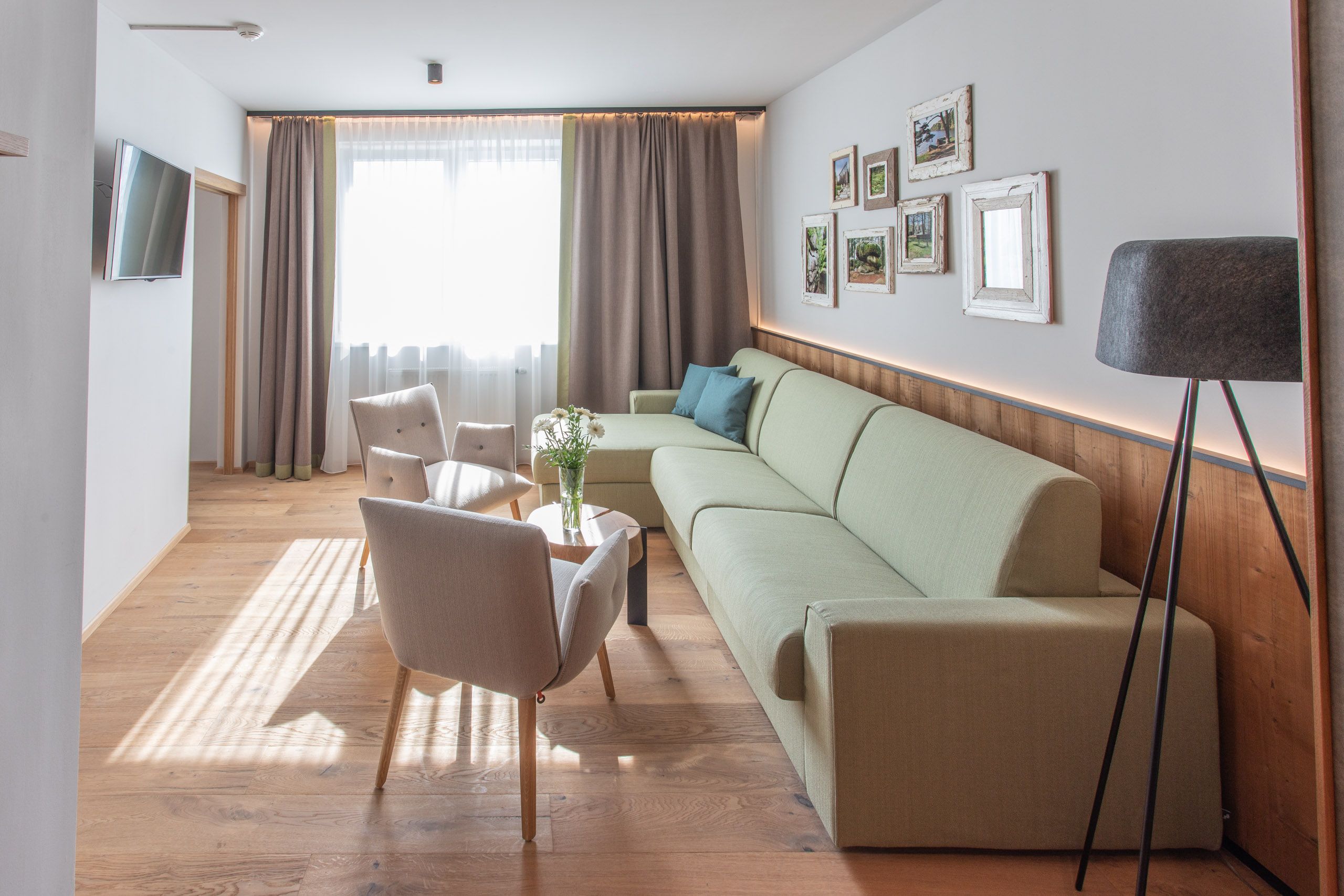 Helles Hotelzimmer mit grüner Couch, zwei Sesseln, Holzboden und Wandbildern.