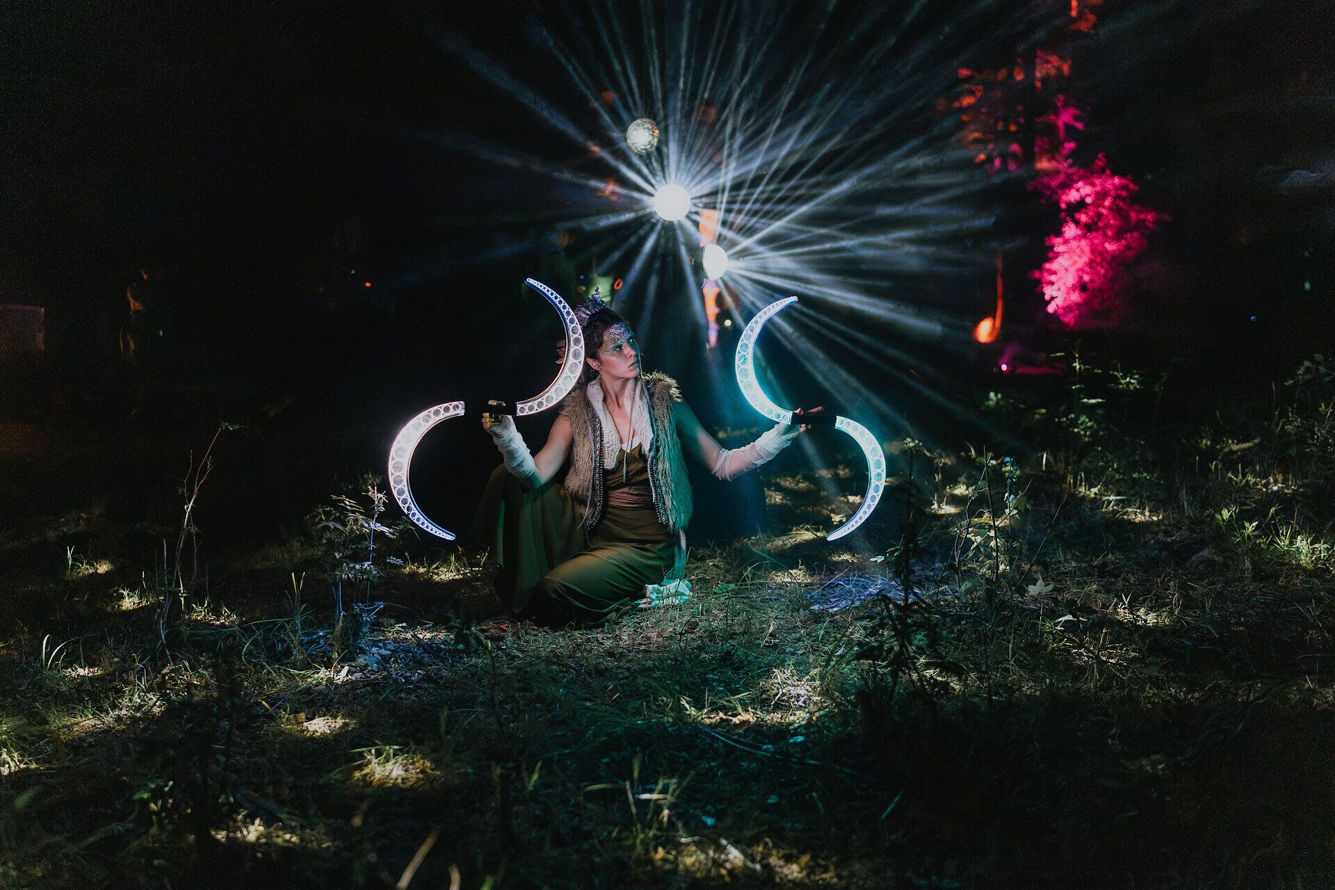Nächtliche Performance im Wald: Eine Person kniet auf dem Boden und hält leuchtende, halbmondförmige Lichtobjekte in den Händen; hinter ihr strahlen Lichtkegel durch den Wald, Bäume sind farbig illuminiert.